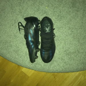 Svarta adidas fotbollsskor predators strl 41⅓  - Säljer ett par svarta adidas fotbollsskor med blå detaljer. Skorna har snörning och mönstrad sula med dobbar för bra grepp på planen. Tillverkade i syntetmaterial och levereras i originalkartong. Perfekta för dig som vill ha stilrena och snabba skor på matchen.