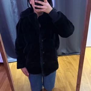 Svart pälsjacka från Millenium, S - Mysig svart pälsjacka från Millenium i storlek S. Jackan har en fluffig känsla, klassisk krage och knäpps med knappar framtill. Perfekt för dig som vill ha en stilren och varm look under kyliga dagar.