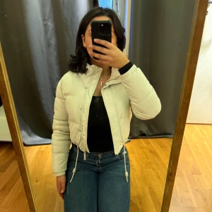 Vit pufferjacka från FB Sister XS - Säljer en snygg vit pufferjacka från FB Sister i storlek XS. Jackan är croppad med dragsko i midjan, hög krage och dragkedja framtill. Perfekt för kalla dagar och ger en clean look till alla outfits.