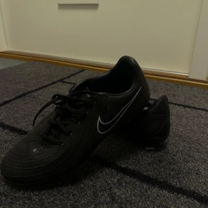 Svarta Nike fotbollsskor med dobbar - Säljer ett par svarta Nike fotbollsskor med klassisk vit swoosh på sidan. Skorna har snörning och rund tå, samt dobbar undertill för bra grepp på planen. Tillverkade i syntetmaterial med diskret mönster på ovandelen för extra stilpoäng.