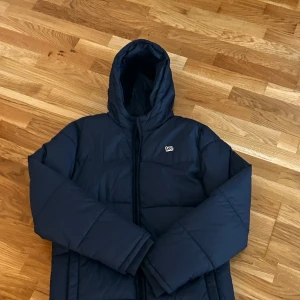 Mörkblå pufferjacka från Lee - Säljer en mörkblå pufferjacka från Lee med huva och dragkedja. Jackan är vadderad och har en stilren look med diskret logga på bröstet. Perfekt för kalla dagar när du vill hålla värmen och ändå se snygg ut.