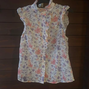 Blommig blus från Holly & Whyte - Supersöt blommig blus från Holly & Whyte i vitt med lila och korallfärgade blommor. Blusen har volangkant längs knäppningen, liten ståkrage och är ärmlös med rynkade axlar. Perfekt för vår och sommar när du vill ha något luftigt och färgglatt.