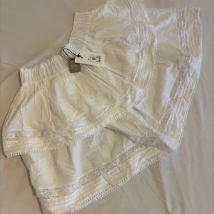 Vit volangkjol med spetsdetaljer - Superfina vita shorts från BikBok med bred resår i midjan, volangig form och dekorativa spetsdetaljer längs kanterna. Lätta och luftiga i bomull, perfekta för varma dagar. Prislapp på och aldrig använd.