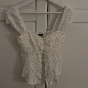 Vit blus med knappar från Gina Tricot - Supersöt vit blus från Gina Tricot med smockad struktur, breda axelband och dekorativa knappar framtill. Toppen har en lätt volangkant nertill och är tillverkad i ett mjukt bomullsliknande material. Perfekt för en fräsch och somrig look!