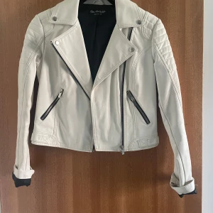 Vit skinnjacka med dragkedjor - Snygg vit skinnjacka från Blue Soul Style med coola detaljer som snedställda dragkedjor, quiltade axlar och spännen vid ärmslut och midja. Jackan har en klassisk bikerstil och är figursydd med lång ärm och svart innerfoder.