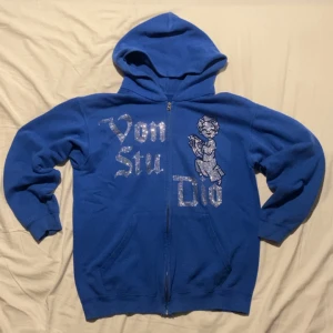 Blå Von Studio hoodie  - Blå hoodie från Von Studio med glittrande strassdetaljer och en stor ängel på framsidan. Dragkedja hela vägen, huva och två fickor. Perfekt statement-plagg för dig som gillar att sticka ut.