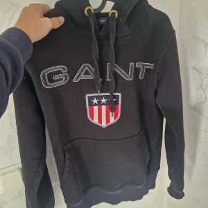 Svart hoodie från Gant med logga - Svart hoodie från Gant med broderad logga och sköld på bröstet. Tröjan har huva med snörning, känguruficka och ribbade muddar. Perfekt för dig som gillar klassisk streetstyle och vill ha något bekvämt och snyggt.