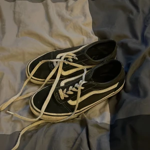 Svarta Vans Old Skool sneakers - Klassiska Vans Old Skool sneakers i svart med vit sula och vit sidostrip. Skorna har snörning och är tillverkade i canvas och skinn. Perfekta för dig som gillar streetstyle och vill ha en tidlös look.