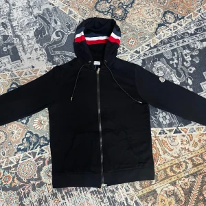 Svart Moncler kofta med dragkedja - Svart kofta från Moncler med hel dragkedja, huva med blå, vit och röd rand samt matchande detalj på ärmslutet. Klassisk Moncler-logga på ärmen och känguruficka framtill. Tillverkad i mjuk bomull, perfekt för säsongens alla chill dagar.