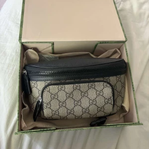 Gucci beige magväska med GG-mönster - Snygg magväska från Gucci i beige canvas med klassiskt GG-mönster och svarta skinn-detaljer. Väskan har två fack med dragkedja och justerbart band. Perfekt för att bära det viktigaste och ge din outfit en lyxig touch.