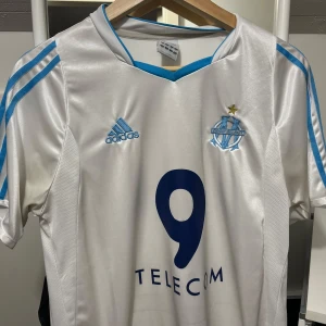 Retro marseille tröja - Säljer en vit Olympique de Marseille fotbollströja från Adidas med ljusblå detaljer och klubbmärke broderat på bröstet. Tröjan har nummer 9 och TELECOM-tryck på framsidan samt klassiska Adidas-ränder på axlarna. Tillverkad i glansigt, ventilerande material. 
