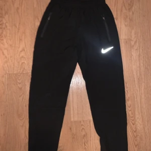 Svarta träningsbyxor från Nike - Svarta träningsbyxor från Nike med elastisk midja och dragkedjefickor. Byxorna har en vit Nike-logga på benet och är tillverkade i ett lätt syntetmaterial som passar perfekt för sport och aktivitet.