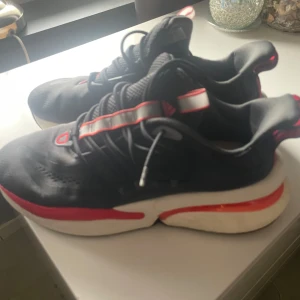 Svarta Adidas sneakers med röd detalj - Snygga svarta Adidas sneakers med röda och vita detaljer. Ovandelen är i mesh och syntet, med snörning och en tjock vit sula med röd accent. Perfekta för dig som gillar sportig stil och vill ha bekväma skor till vardags.