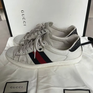 Gucci Ace  - Gucci Ace sneakers i vitt skinn med klassiska röd- och blåa ränder på sidan. Baksidan har svart ormskinnsmönstrad detalj med Gucci-logga. Platt sula, snörning och rund tå. Skorna har slitage o litet hål och därav priset men har mycket kvar att ge. Perfekta för dig som vill ha en ikonisk och stilren sneaker med lyxig känsla. Kvitto finns och skorna är självklart äkta✅