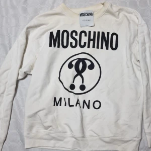 Vit sweatshirt från Moschino - Säljer en vit sweatshirt från Moschino med stor svart logga och Milano-tryck framtill. Tröjan har rund halsringning och långa ärmar. Perfekt för dig som gillar statement-plagg och italiensk design.