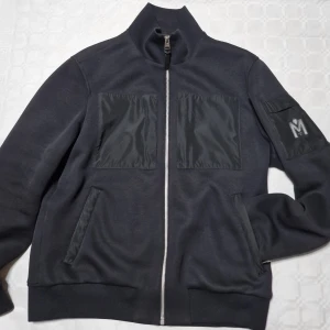 Svart bomberjacka från Mackage - Svart bomberjacka från Mackage med hög krage, hel dragkedja och två stora fickor framtill i blankt material. Jackan har en snygg patch med M-logga på ärmen och ribbade muddar. Perfekt för dig som gillar stilren och modern look.