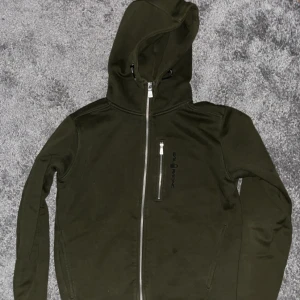 Olivgrön hoodie från Sail Racing - Olivgrön hoodie från Sail Racing med hel dragkedja och huva. Tröjan har en bröstficka med dragkedja och diskret logga på bröstet. Mjuk insida och långa ärmar, perfekt för kyliga dagar.