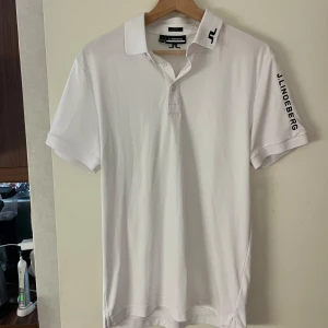 Vit piké från J.Lindeberg - Vit pikétröja från J.Lindeberg i nyskick med svart logga på kragen och ärmen. Klassisk krage med knappar och korta ärmar. Tillverkad i mjuk bomull, perfekt för en clean och sportig look.