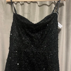 Svart glittrig långklänning med paljetter  - Supersnygg svart långklänning med tunna axelband och massor av glittrande paljetter. Klänningen har en elegant draperad urringning och är perfekt för dig som vill sticka ut. Den är figurnära upptill och faller snyggt ner mot golvet. 