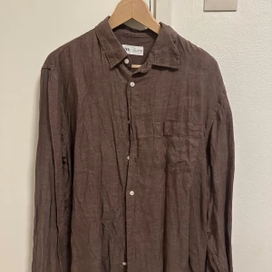 Brun linneskjorta från Zara, L - Snygg brun skjorta från Zara i storlek L, tillverkad i svalt linne. Skjortan har klassisk krage, bröstficka och vita knappar framtill. Perfekt för en avslappnad och stilren look.