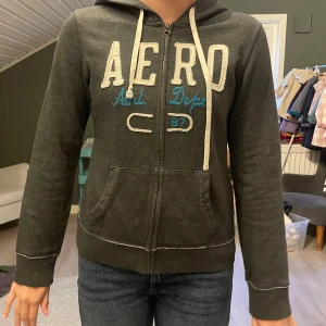 Mörkgrå hoodie från Aeropostale - Mörkgrå hoodie från Aeropostale med vit broderad logga och blå detaljer framtill. Tröjan har dragkedja, huva med snörning och två fickor framtill. Står storlek L men känns mer som S/M