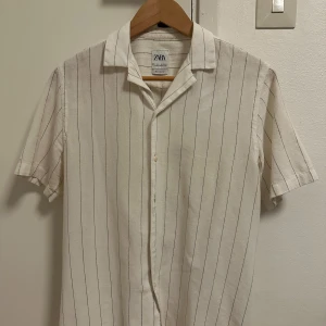 Vit randig skjorta från Zara - Vit skjorta från Zara i relaxed fit med tunna svarta ränder. Skjortan har korta ärmar, klassisk krage och knäppning framtill. Perfekt för en chill och stilren look. Tillverkad i ett mjukt bomullsmaterial.