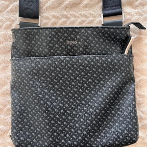 Hugo Boss Axelväska - Snygg svart axelväska från BOSS med diskret grått rutmönster och justerbar axelrem med logga. Väskan har dragkedja upptill, innerfack med dragkedja och är tillverkad i slitstark syntet. Perfekt för dig som vill ha en stilren och praktisk väska med premiumkänsla.