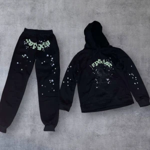 Svart Sp5der  - Svart hoodie med huva och snörning, dekorerad med stjärnor på ärmarna och ett  grönt Sp5der tryck framtill. Materialet är mjukt och skönt, perfekt för chill dagar. Matchande svarta byxor med samma stjärnmönster ingår.