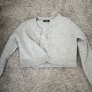 Grå croppad kofta från FB Sister - Mysig ljusgrå croppad kofta från FB Sister med rund halsringning och knappar framtill. Stickad i mjukt material och har långa ärmar samt ribbade muddar vid ärmslut och nederkant. Perfekt för lager-på-lager och chill dagar.