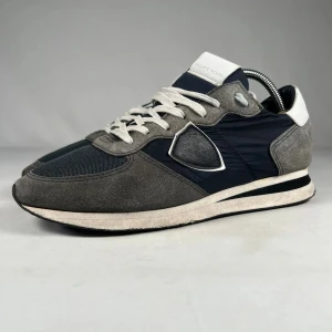 Philippe Model sneakers i grå och svart - Snygga sneakers från Philippe Model i en mix av grå mocka och svart  textil. Skorna har vita detaljer vid häl och plös, samt vita snören. Klassisk sportig siluett med rund tå och platt sula. Perfekta för dig som gillar stilrena och bekväma sneakers.