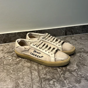 Saint Laurent vita sneakers med text - Unika vita sneakers från Saint Laurent. Priset kan ändras. Skorna har slitna detaljer, beige sula och klassisk snörning. Tillverkade i canvas med inslag av skinn vid hälen. Perfekta för dig som gillar en edgy och avslappnad stil.