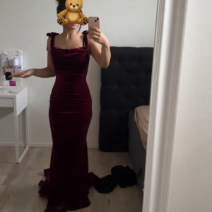 Vinröd sammetsklänning med rosettband - Supersnygg långklänning i vinröd sammet med figurnära passform. Klänningen har breda axelband med rosettknutar och en elegant, lätt utsvängd nederdel. Perfekt för dig som vill sticka ut med en lyxig och trendig look.