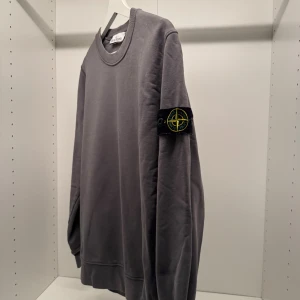 Grå Stone Island sweatshirt - Säljer en grå sweatshirt från Stone Island med klassisk rund hals och ikonisk patch på vänster ärm. Tröjan har en clean look, ribbade muddar och är tillverkad i mjuk bomull. Perfekt för dig som gillar streetwear och vill ha något stilrent.
