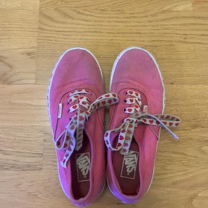 Rosa Vans sneakers med hjärtan - Är inte i så bra skick men går att tvätta på vissa ställen. Säljer ett par rosa Vans sneakers i tyg med vita sulor dekorerade med röda hjärtan. Skosnörena är grå med röda hjärtan, vilket ger en gullig och unik look. Klassisk rund tå och platt sula, perfekta för dig som vill sticka ut lite extra.