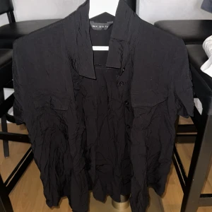 Svart kortärmad skjorta, strl M - Svart skjorta med korta ärmar och klassisk krage. Skjortan har knappar framtill och två bröstfickor med lock. Materialet är lätt och skrynklar sig snyggt för en avslappnad look. Perfekt till jeans eller shorts för en chill stil.