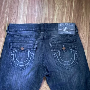 Svarta True Religion jeans - Säljer ett par svarta jeans från True Religion med ikoniska bakfickor och kontrastsömmar. Jeansen har snygga slitningar framtill och raka ben. Klassisk femficksmodell med knappstängning och detaljerade ficklock bak.