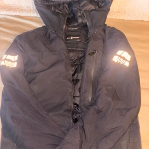 Svart jacka Sail Racing - Svart pufferjacka från Sail Racing med huva och reflekterande loggor på båda ärmarna. Jackan har dragkedja framtill och en bröstficka med dragkedja. Perfekt för kalla dagar.