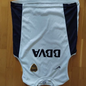 Vit ärmlös Boca Juniors fotbollströja - Säljer en vit ärmlös Boca Juniors fotbollströja från Nike med svarta sidopaneler, BBVA-tryck på bröstet och klubbmärke i gult och svart. Perfekt för träning eller match. Materialet är lätt och andas bra.