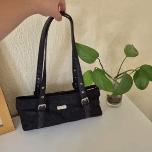 Svart handväska från Dolce & Gabbana - Stilren svart handväska från Dolce & Gabbana med silverfärgad logga framtill. Väskan har långa handtag med snygga spänndetaljer och dragkedja upptill. Utsidan är i textil med diskreta logotypdetaljer och insidan har ett brunt foder med logotypmönster. Använd ett fåtal gånger, mycket bra skick!!