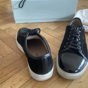 Svarta sneakers från Lanvin i mocka - Snygga svarta sneakers från Lanvin med ovandel i mocka och lackad tå. Skorna har vita platta sulor, svarta snören och rund tå. Kommer med originalkartong och dustbags. Perfekta för dig som gillar stilrena och lyxiga detaljer. Lanvin skor dm för info