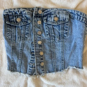 Jeans tubtopp med knappar och fickor - Säljer en snygg tubtopp i ljusblå denim med silverfärgade knappar framtill och två bröstfickor. Toppen har fransig nederkant och smockad rygg för skön passform. Perfekt för dig som vill ha en cool och trendig look. Har ingen prislapp men tror den är xs