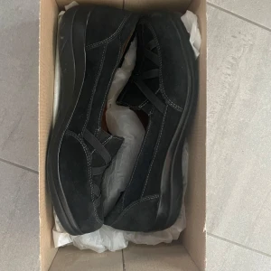 Svarta mockaloafers från Natural Soul - Svarta loafers i mocka från Natural Soul med dekorativa korsade band framtill och kontrastsömmar. Skorna har en rund tå och platt sula, samt en mjuk innersula för extra komfort. Perfekta för dig som gillar en stilren och bekväm look.