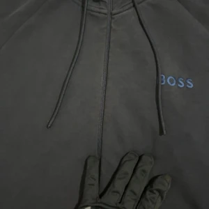 Mörkblå hoodie från BOSS med dragkedja - Svart hoodie från BOSS med broderad logga på bröstet och snygg zip-dragkedja. Tröjan har snörning i huvan och praktiska fickor framtill. Tillverkad i mjuk bomullsmix, perfekt för chill eller streetwear.