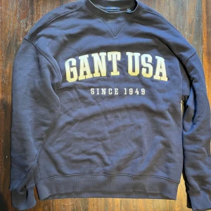 Mörkblå sweatshirt från Gant - Säljer en mörkblå sweatshirt från Gant med stor gul och vit logga 'GANT USA' och texten 'Since 1949' på bröstet. Tröjan har rund halsringning, långa ärmar och ribbade muddar. Perfekt för en chill och sportig look. 