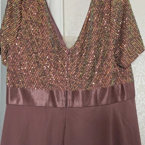 Glittrig långklänning i brons - Långklänning i bronsig ton med glittriga paljetter på överdelen och djup v-ringning. Överdelen har diagonala paljettmönster i guld, grönt och koppar, samt bred satinliknande midja. Kjolen är enkel och faller snyggt. Perfekt för dig som vill sticka ut.