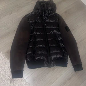 Mackage Cardigan - Snygg svart Cardigan från Mackage med glansig vadderad framsida och huva. Jag säljer denna Cardiganen för 750 eftersom den är solbränd men det går att ha på sig ändå. Du kan skicka den till en slags företag som fixar det solbrända
