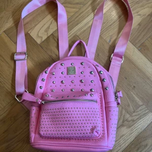 Rosa ryggsäck med nitar - Cool rosa ryggsäck från MAM med silvriga nitar och dragkedja. Väskan är gjord i syntetmaterial med struktur och har justerbara axelband. Perfekt för dig som vill sticka ut med en färgglad och edgy accessoar.