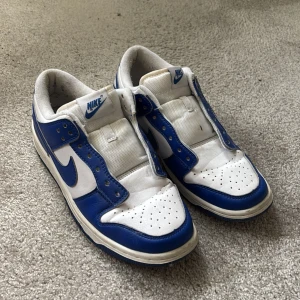 Nike Dunk Low blå och vita sneakers - Nike Dunk Low sneakers i blått och vitt med klassisk swoosh på sidan. Skorna har perforerad tå, platt sula och rund tå. Ovandelen är i skinn och textil, med blå yttersula och vit/blå färgblockning. Nike-logga på plös och häl. Perfekt för dig som gillar retrostil. Utan skosnören . Stl 42 ( 26,5cm)