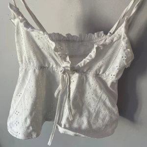 Vit broderad linnetopp från SHEIN - Söt vit linnetopp från SHEIN i storlek XS. Toppen har smala axelband, v-ringning med volangkant och knyt framtill. Broderade detaljer och lätt struktur i tyget ger en romantisk känsla. Perfekt för varma dagar och somriga outfits.
