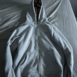 Grå hoodie från Burberry med dragkedja - Snygg grå hoodie från Burberry med klassiskt rutigt foder i huvan. Tröjan har hel dragkedja, två fickor fram och justerbar huva med snörning. Perfekt för dig som vill ha en stilren och lyxig vardagslook.